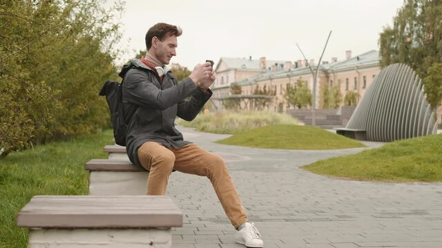 Full shot of happy Caucasian man dressed casually, sitting on small bench in public square, taking pictures with smartphone of invisible object, smiling, checking photograph afterwards