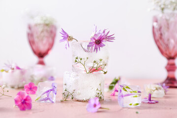 Glasses with beautiful flowers frozen in ice on color background