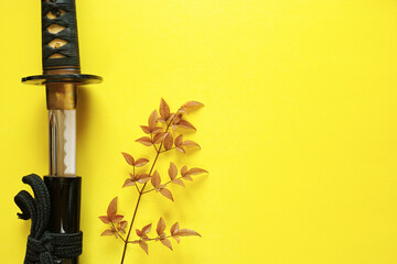 Fototapeta na wymiar 黄色い紙の背景の左に置いた抜きかけの日本刀と赤い葉の木の枝。右側半分がコピースペース。