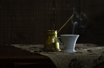 White cup with hot steaming coffee and cezve on a dark background.
