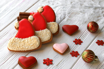 Christmas cookies, gingerbread cookies in form of red Santa Claus cap and heart with icing lie among the cinnamon, New Year decorations on white wooden background. Christmas food 2021