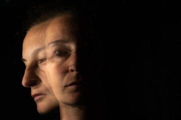 Dark portrait of a serious woman with only half her face lit up on a black background looking sad or in a depression. The face unfolds into another blurred and equally sad face.