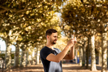 Young man in tshirt standing on street and makes photo on mobile phone. In the background is a tree lane.