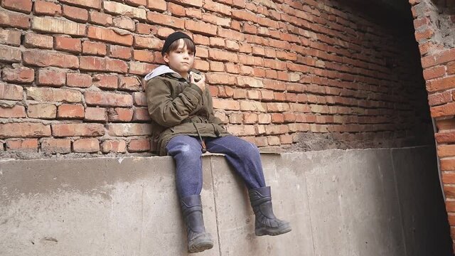 Street kid boy sits on the concrete outdoors looking at camera, having no home, need shelter.