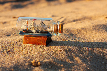 Horizontal close-up photo of a miniature souvenir of a vintage three-masted sailing ship (barque) in a small transparent glass bottle, closed with a cork, on the sand during sunset