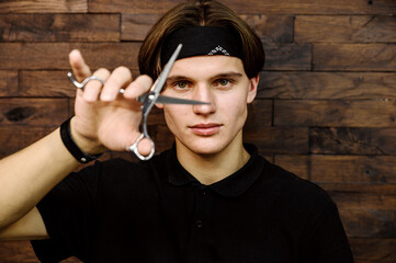 portrait of stylish barbershop master on dark wooden background. professional young hairdresser in black bandage holds silver scissors near the face.