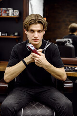 stylish and young master of haircuts sits in a wrestle holding tools for a haircut. scissors and a typewriter in the hands of a barber. barbershop and a portrait of the master. beauty salon