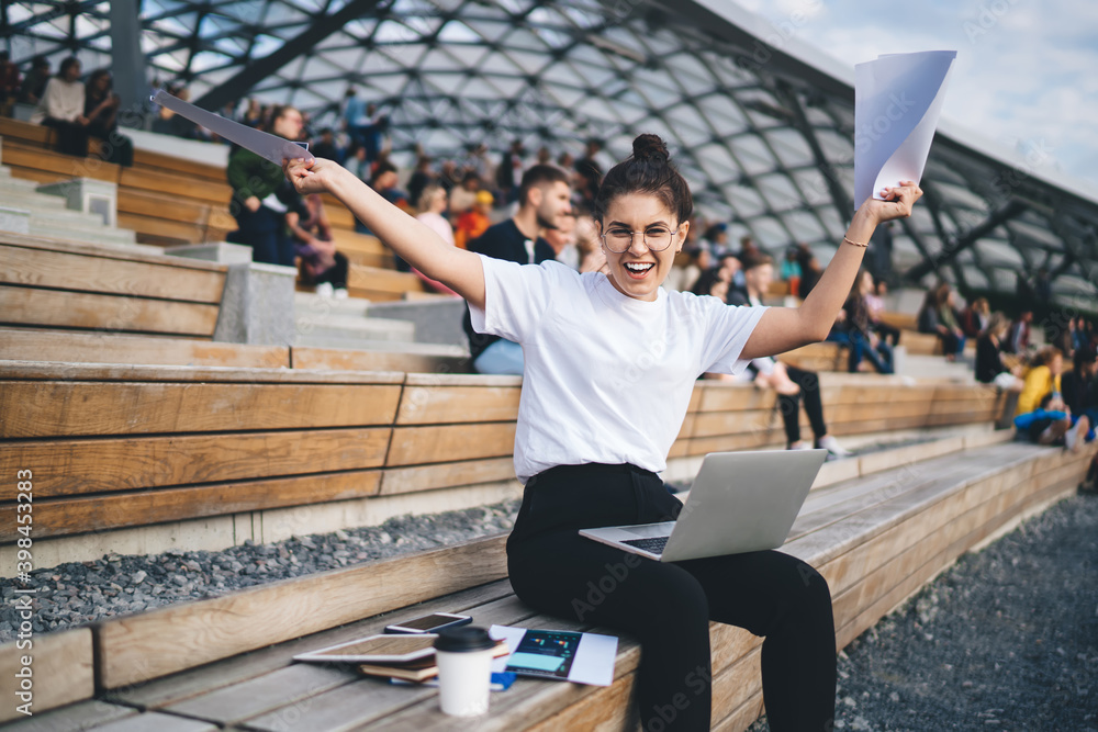 Poster Portrait of exited female freelancer enjoying time for remote working with documents and modern laptop computer, happy Caucasian student rejoicing in public place using netbook and papers for planning