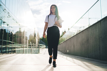 Youthful hipster youngster dressed in trendy casual clothing holding education equipment for learning and walking at campus urbanity, Caucasian woman with modern touch pad smiling at city street