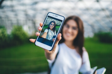 Selective focus on smartphone screen with displayed selfie image on it, blurred hipster girl shooting influence video during leisure time for creating multimedia content via cellphone application