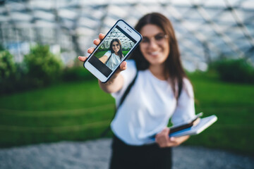 Selective focus on smartphone screen with displayed selfie image on it, blurred hipster girl shooting influence video during leisure time for creating multimedia content via cellphone application