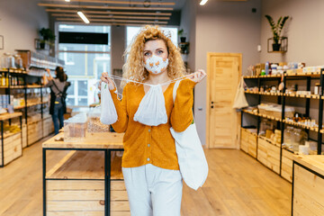 Blond curly woman in reusable protective face mask using textile bag on plastic free grocery store on background. Eco friendly shopper bag in female's hands