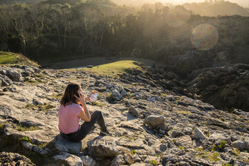 Young woman sitting over a rock in the nature during sunset. She is making a phone call and holding a sandwich.