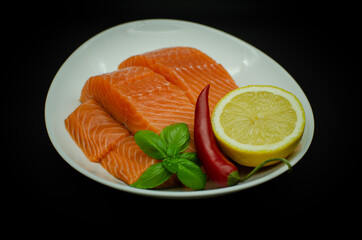 Pieces of Fresh Salmon fillet laying on white plate. isolated on black. Served with chilli pepper, lemon and greens