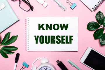 Fototapeta na wymiar KNOW YOURSELF is written in a white notebook on a pink background surrounded by business accessories and green leaves.