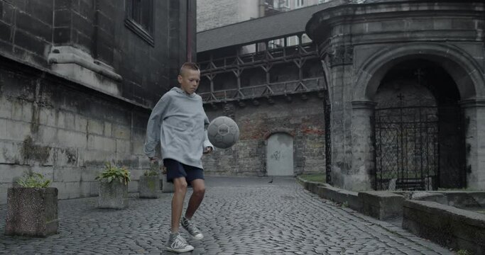 Capable teenager guy kicking football ball with feet . Young talanted boy s practicing tricks at old european city street. Concept of sport, talent and lifestyle