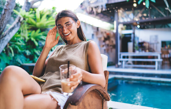 Joyful woman with drink sitting near swimming pool