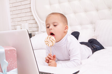 A boy with Down syndrome is eating a candy on a stick and looking at a laptop, a video call and...