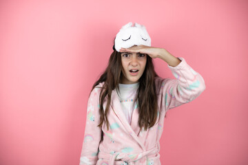 Pretty girl wearing pajamas and sleep mask over pink background looking far away with hand over head. Searching concept.