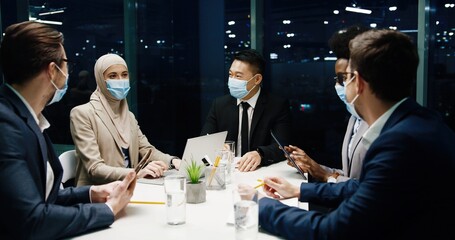 Happy multi-ethnic male and female proessionals in medical masks in modern office having team business conference working in evening and speaking about business startup, typing on laptop and tablet