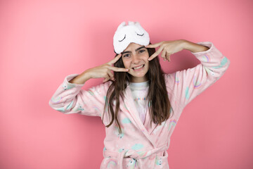 Pretty girl wearing pajamas and sleep mask over pink background Doing peace symbol with fingers over face, smiling cheerful showing victory