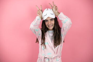 Pretty girl wearing pajamas and sleep mask over pink background Posing funny and crazy with fingers on head as bunny ears, smiling cheerful