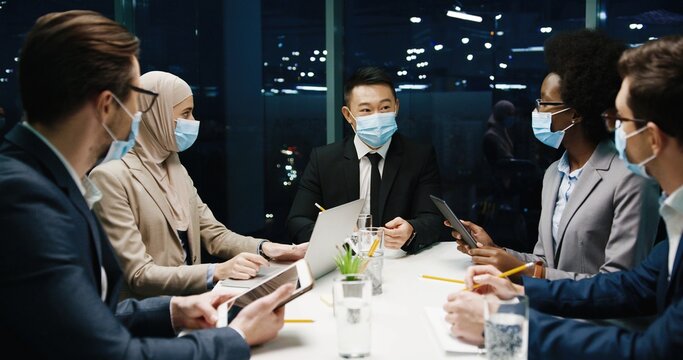 Happy multi-ethnic male and female proessionals in medical masks in modern office having team business conference working in evening and speaking about business startup, typing on laptop and tablet