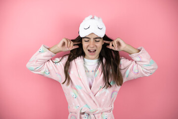 Pretty girl wearing pajamas and sleep mask over pink background covering ears with fingers with annoyed expression for the noise of loud music. Deaf concept.