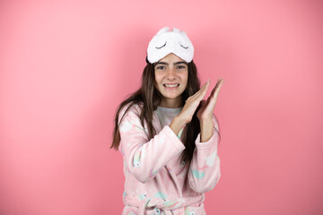 Fototapeta premium Pretty girl wearing pajamas and sleep mask over pink background clapping and applauding happy and joyful, smiling proud hands together