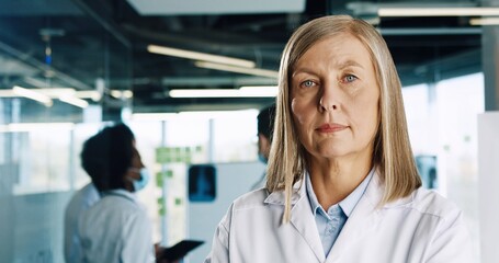 Close up portrait of elderly Caucasian woman doctor wearing white coat standing in clinic hall and looking at camera. Mixed-races doctors in masks working on background. Coronavirus concept