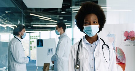 Close up portrait of young african American woman doctor wearing protective medical mask standing in clinic and looking at camera. Caucasian doctors working on background. Coronavirus concept