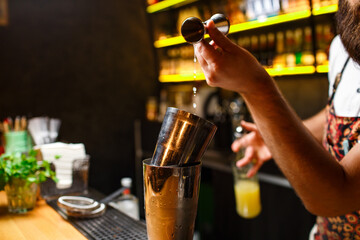 bartender is preparing a cocktail in a shaker. bartender's hand adds prescription ingredient to steel shaker on dark background highlighted by yellow bar