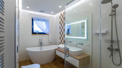 Fototapeta na wymiar Contemporary interior of bathroom in luxary apartment. Double sink on countertop. White toilet and bathtub. Illuminated mirror. Shower.