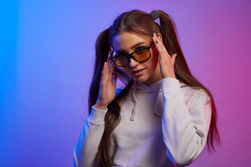 Portrait of young woman wearing trendy tinted yellow lens sunglasses seriously looking straight on camera in neon light. Hipster teenager in glasses.