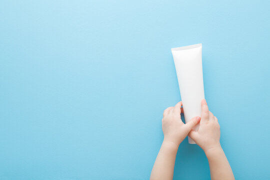 Baby little hands holding white cream tube on light pastel blue table background. Care about clean and soft body skin. Children beauty product. Empty place for text. Top down view. Copy space. 