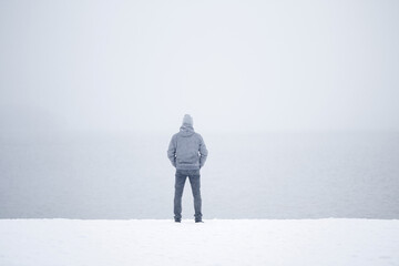 Young adult man standing alone on snow at lake shore and looking far away. White cold snowy winter day. Thinking about life. Mist over water. Peaceful atmosphere in nature. Back view.