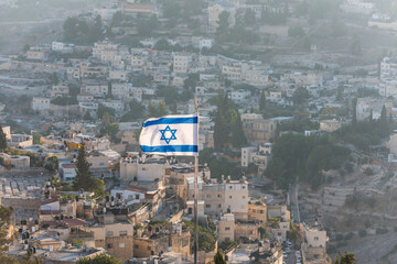 Israeli National flag waving on the top of Mount of Olive with background of residential houses in...