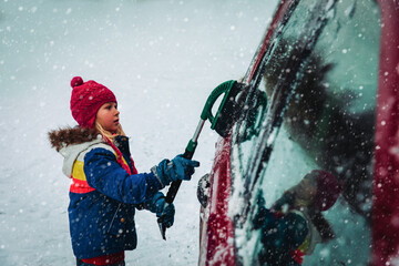 Child brushing snow off car. Kid with winter brush and scraper clearing family car.