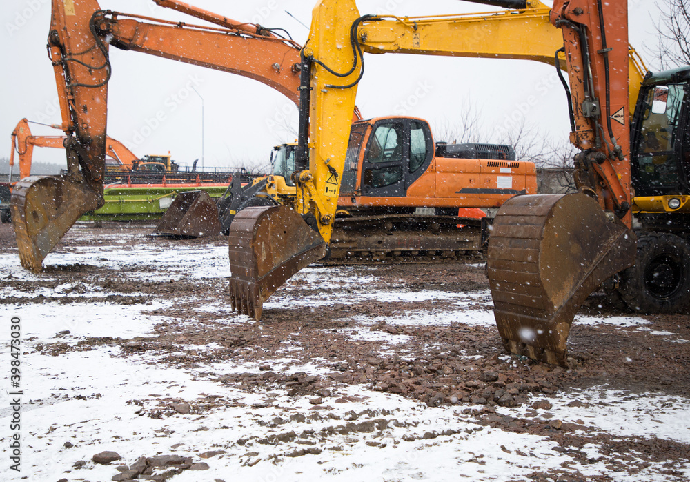 Wall mural large orange and yellow used crawler excavators standing side by side at a construction site. Snowing, winter. Three iron buckets in row