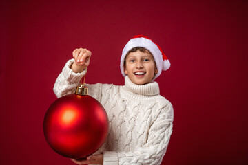 happy boy in Santa hat and knitted sweater holds Christmas ball in his hands