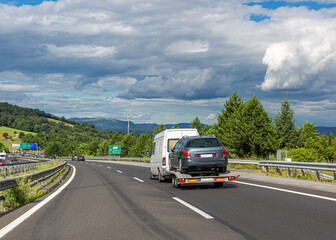 White minibus with tow truck transporter on highway. Car carrier trailer with used car. Beautiful landscape background