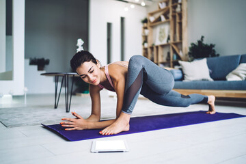 Fototapeta na wymiar Strong female in casual sportive wear standing in asana on carpet stretching body keeping muscular strength, fit girl enjoying morning yoga for training flexibility in modern home interior