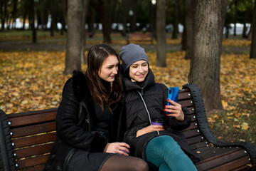 Fototapeta na wymiar Two girls in the park talk on a bench with a phone and a glass.