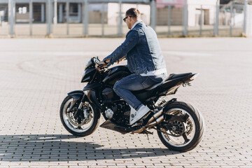 Obraz na płótnie Canvas A man in denim clothes sitting on a black sports motorcycle against the backdrop of a large modern building