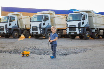 cute boy plays with a toy dump truck on control panel on asphalt. behind him are large real dump trucks. sunny warm day. Favorite games for boys © Anna