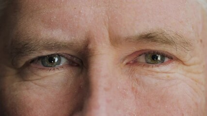 A friendly and flirty elderly mature man winking with blue eyes. A wink by middle aged male senior citizen with wrinkles on attractive face looking at camera. Slow motion, macro extreme close up, 4K.