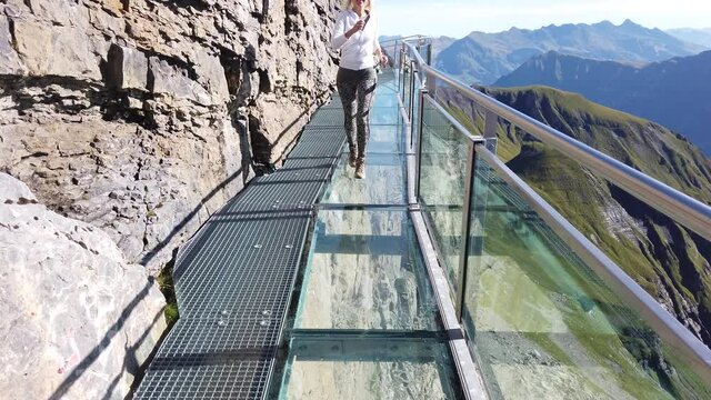 Outdoor activities in Birg, 2677, on Schilthorn in Bernese Prealps, Canton of Bern, Switzerland. Happy tourist woman walks the Thrill walk on a crystal floor suspended above village of Murren.