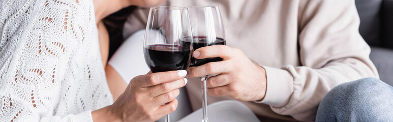 Cropped view of glasses of red wine in hands of young couple on blurred background, banner