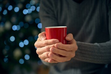 A man holds a red cup of tea or hot chocolate in his hands to warm himself from the cold of winter. In the background, the lights create a bokeh effect to celebrate holy holidays and warm the room.