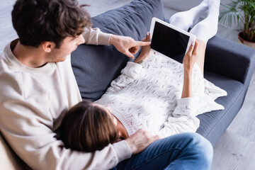 Man pointing at digital tablet with blank screen near girlfriend on couch on blurred foreground